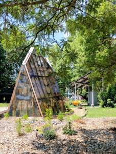 “Shine A Light” is solar-powered triangular pod-like structure, with a structure of cedar boards covered with a semi translucent sheath with patterns on the surface like oversized wood grain. The structure is set in a garden with cedar mulch and overhanging trees, and a solar array is behind the triangular structure to the right.