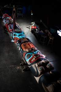 “Kaagwaantaan Kooteeyaa” is a large totem pole lying diagonally in a dark studio area. Various animal and human faces are sculpted in wood, primarily painted in red, turquoise and black.