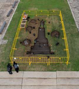 “Shadow on the land, an excavation and bush burial” is a sculptural installation in a grass park square. Yellow crowd control barriers frame an excavation site, and at the center, sod has been removed to create a silhouette of a figure on top of a monumental pedestal. Staked string frames various areas, and tools and buckets dot the area.