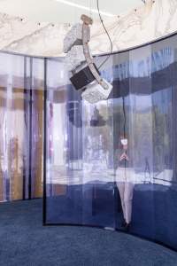 “the input of this machine is the power an output contains” is a multimedia installation, in which a performer is reciting a text generated by artificial intelligence. In this view, a masked person stands behind a blueish curved plexiglass wall, and from the ceiling hangs a curved array of 3 cinder blocks covered with fiber on the face and a black speaker of the same size.