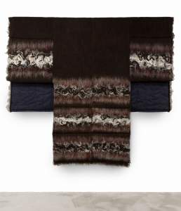 “Sound Blanket #6” is a fiber sculpture hanging on a white wall, in the form of a flat hanging kimono formed from bands of dark bands of dark wool, felted swirls of white, gray, and black wool and hair, and dark denim.