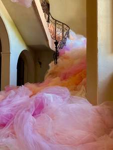 “Déferlante” is a sculptural installation in a grand curving staircase with an iron filigree balustrade framed by warm stucco walls. Tulle in hues of pink, peach, and fuschia fill and cascade down the staircase, filling the landing below.