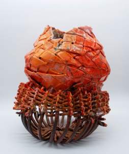“Crawfish Armor” is a stoneware structure, with a base of brown curved rods in a cup form, woven through at the top with red-orange wishbone stacks. Sitting on top are ceramic strips woven into a rounded form with an open top. The color shifts from deep to light orange with dark speckles and drips.