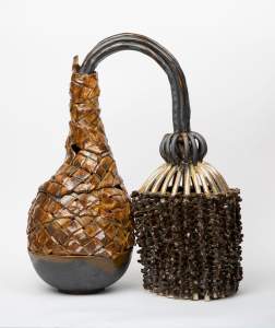 “Bridged Over” is a stoneware sculpture of two connected vessels. On the left is a vase-like form of brown glazed woven strips extending from a bowl. Tubes of gathered black ceramic tubes arc from the mouth of the vase, and bubble out to create a sphere and cylindrical cage, woven on the sides with glossy dark brown pinched clay.