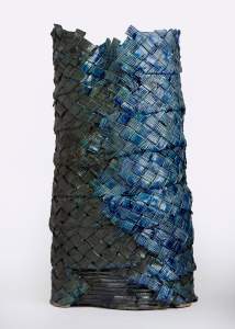 “Chasing Waterfalls” is a stoneware structure of basket-woven ribbed strips of ceramic, wrapped in bands to form an irregular vertical cylinder. The shiny glaze transitions from olive green to blue and turquoise.
