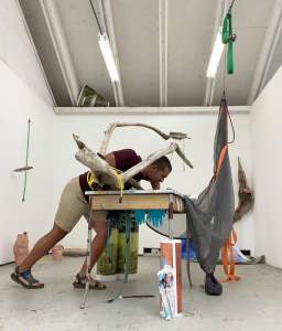 In this performance of “Losing my face in the lake” Javier Orfon leans over a distressed industrial desk under a gnarled piece of driftwood. Blue rubber gloves, and colored elastic cords hang from the bottom of the desk, and digital prints of landscapes stand in coiled cylinders around one desk leg and behind him. A gray bug net drapes off a corner of the desk, and is partially suspended from the ceiling with a tangle of orange cording.