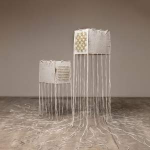 “Visitation” is a sculpture of 2 rectangular wide cubes made of textured off white handmade paper floating next to each other, bound at the edges with tied white deer hide laces. Long strings of lacing hang down from the edges and pool on the floor. The front cube is higher up in the air, and on one face has a checkerboard pattern of tan porcupine quills protruding an inch from the surface. The front face of the rear cube has 6 horizontal lines of quills protruding.