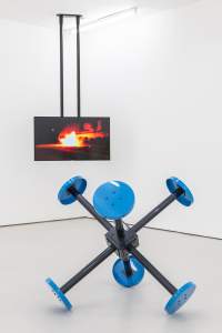 “PIVOT I” is a sculptural installation in a white room. In the foreground, 6 1:1 scale replicas of prison visiting room stools with blue tops and black stems are fused together at the base like a large pronged metal Jack toy. In the background, a tv screen hangs from the ceiling displaying an image of a vehicle burning on a street at night.