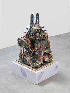 Big Dog (Guardian) is a colorful, abstract ceramic sculpture resembling an anthropomorphic coyote-like creature crouched and decorated with many fine details, holding a receptacle with a small human figure inside.