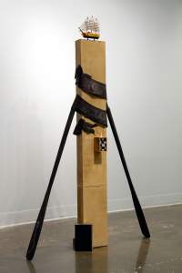 Don’t hate the player, hate the Game is a sculpture centered on a tall rectangular column with a model ship at the top. Two wooden oars extend from the sides, and a black banner encircles the column. A pawn chess piece stands on a small box protruding from the lower center, and a book leans at the foot of the column.