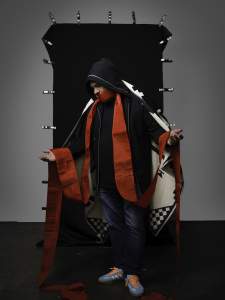 In Mourning the Motherland (Praying) the artist stands in front of a clamped black backdrop with outstretched open hands and bent head. He wears a patterned cloak, black hood, and a strip of red fabric covers his mouth and drapes down his lapels and over his hands.