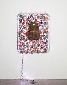 Unattended Baggage (Modern Warfare) is an installation with camo backpack with an LED countdown reading 00:00:02:50, hanging in a metal frame surrounded by various embroidery patches, and string lights which are also  entwined around the frame and extending out to a plug on the floor.