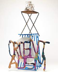 Untitled (Orientalism charm with TV tray) is a mixed media assemblage structure. At the base, sculptural words read “ORIENTALISM, in front of a white steel disc, and blue security bars. On top, a TV tray teeters, upon which rests a white sculptural rubble of tangled ceramic, concrete, and wire.