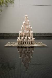 Untitled (15 Esthers in a pyramid) is a sculpture of 15 white poodles made of mottled clay stacked in a cheerleader-like pyramid formation. They stand on a rectangular cement plinth with arranged objects above a dark reflective pool.