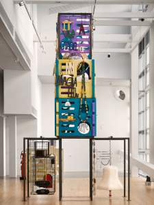 Untitled (radio clock tower with accessories) is a tall, colorful mixed-media sculpture. The base are dark metal frames, from one hangs a large white bell and string of oversized beads, and from the other two animal cages with stereos inside. Tacked about the frame in the center are interlocking plastic squares from children's playhouses in purple, yellow and teal, from which strings of beads and sporadic sculptural text hangs, reading ”It was a citadel of my imagination”. It stands in a bright gallery room with large windows and a high ceiling.