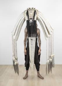 A person stands with bare tattooed arms, wearing a large, elaborate symmetrical headdress with hanging white ropes, long black fringe, and padded sections, obscuring their face and upper body.
