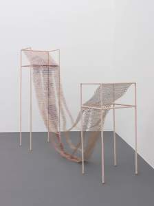 Reclining Nude is a sculpture composed of two thin scaffold-like chairs in different heights, between which is draped a long mesh form of delicate filigree, with its separated strips draping down to touch the floor.