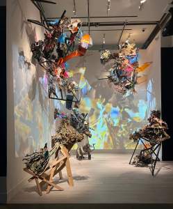 Boru Sibaso Paet, on the foam of the primoridal sea is a mixed media installation in the corner of a darkened gallery. Abstract fragmented shapes are projected on the corner wall, and in front, several abstract assemblage sculptures sit on the floor and others are suspended from the ceiling.