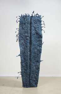 This free-standing, sculptural piece stands like a monolith against a white wall and floor. The sculpture is flat across the top and tapers slightly at the foot, like a coffin. The dark, marine-blue surface is deeply textured with wrinkles and bulges, and drops of the blue material seem to splash out from the edges, especially around the top. Beads line the tendrils leading to the drop-shaped ends.