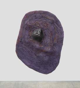 A roughly oval sculptural form hangs on a white gallery wall. The plum-purple surface is wrinkled and deeply textured around a triangular, boulder-like shape near the center.