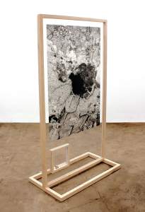 A black-and-white drawing is sandwiched in clear acrylic sheets held in place in a wood-framed floor stand. A bullet and casing are encased in a smaller clear box beneath the drawing, which fills most of the frame. The drawing is gray and veined around an inky black void at its center.