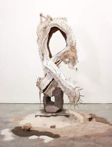 A free-standing sculpture of a serpentine form curves in a loop at the top and cross near the stone on which it perches. The form is covered with white shells, and one leg bristles with metal nails. A deer skull is affixed to the curved section, and the sculpture sits in a pile of sand in different shades of brown.