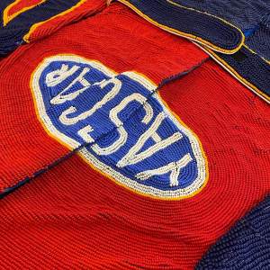 Close up of a suit densely beaded with a logo that reads “YASCAR” in white letters in a bright blue oval. The oval is outlined in white and then yellow against a larger area of crimson red.