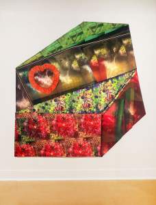Fabric-covered panels on a white gallery wall fit together to create an angular, irregular, six-sided shape. Some panels show scarlet-red flowers, others black and red berries, other plants or landscapes, and patterned cloth. A heart shape patterned with hearts and surrounded by a red ruffle is near the top left corner.
