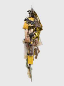 At least two dozen clear plastic pouches hold doll body parts or accessories or materials like shredded gold foil or iridescent strands. Together the hanging pouches create a long sculptural piece that tapers to a point, made of a tube packed with glittery strands, at the bottom.