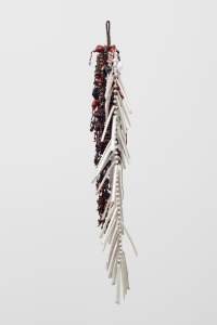 A waterfall-like form of black, brown, and dark red beads makes up about the left half of this long, dangling, sculptural work. White beads and narrow, white, tube-like packets, which are stitched around their edges in white or red, make up the right half. The tubes reach out from the beaded strand like fish bones from a spine.