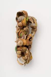 Shards of gourds are arranged in a roughly tall, gourd-like shape and displayed against a white wall. The fragments range from cool peach tones to warm sage green and light tan, and wooden pins bristle across the surfaces.
