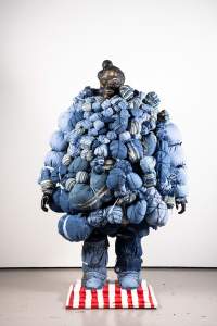 This free-standing sculpture is made up of dozens, perhaps hundreds, of blue denim bundles bound tightly and hung like an oversized coat on a mannequin with a black and gold face. Two black hands hang from the ends of arms also made up of bulging pouches. The legs and feet are covered in layers of blue jeans, and the sculpture stands on a white-and-red striped base in a gallery with a concrete floor and white wall.