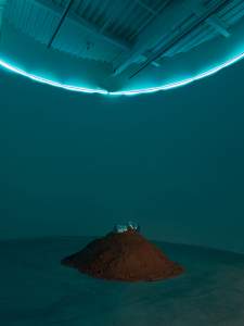 A small model car sits on a mound of dirt in an indoor space, illuminated by a blue neon light above, against a dark teal background.