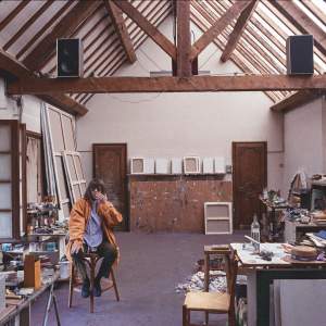 Joan Mitchell sits on a stool in a large studio with a vaulted ceiling with wooden beams and skylights, surrounded with art supplies and canvases of various sizes facing the wall. A white woman with brown hair, she wears an orange raincoat and tinted glasses, and holds a hand to her face.