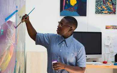 Artist Ashley Teamer, a Black person with dark skin, short cropped black hair, wearing wire frame glasses and a blue button up shirt, paints onto a large print on the wall.