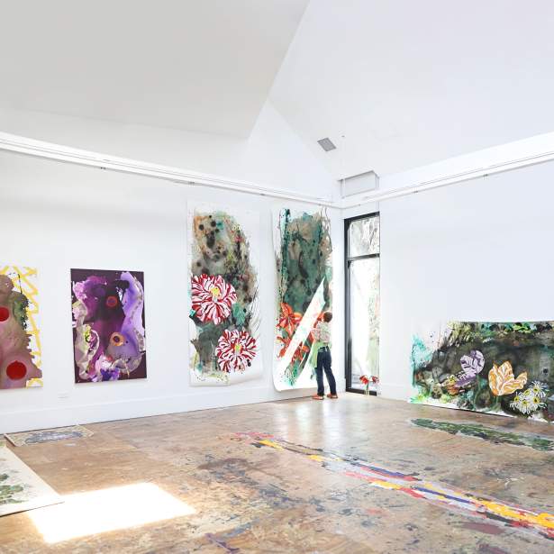 In a large art studio, a white woman with short red hair paints flowers on a large vertical paper sheet. Other large artworks hang, with washes of abstract color, and large flowers.