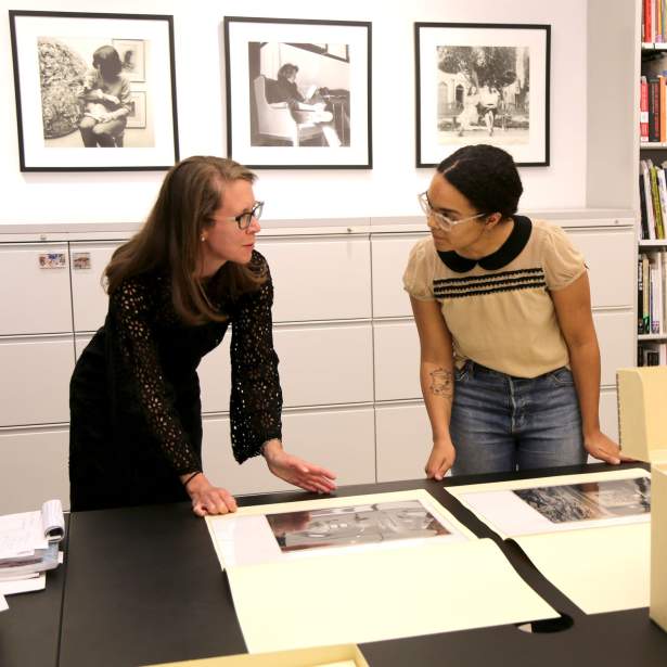Two women talk as they lean over photos in folders.