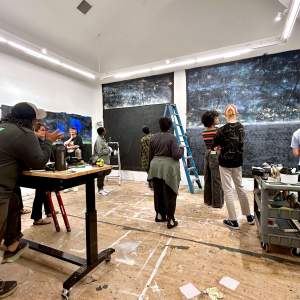 During an Open Studios evening event, several people stand and work in an art studio, surrounded by large abstract paintings, art supplies, and a ladder.