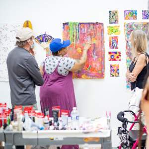 Soraya, with medium dark skin wearing a fuschia dress over a short sleeved patterned tshirt with a royal blue baseball hat, points to a detail of a multicolor abstract work with several guests looking on.