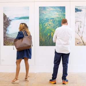 In a white walled studio hang three large vertical photographs of two dreamy seaside landscapes, and an aerial view of green mountains. Facing the artwork are two people: one has medium dark skin and wears a blue dress and wedge sandals, and the other has light skin, wears a white button up, jeans and running shoes.