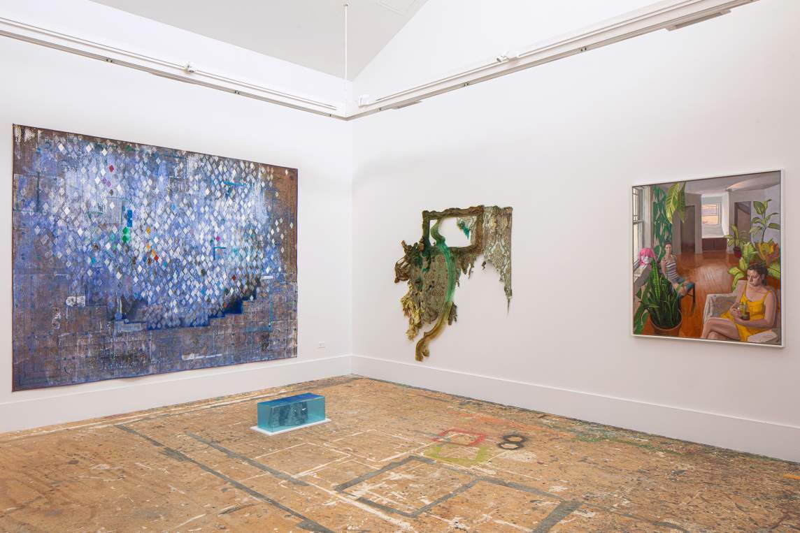 On left, a painting on a patterned rug hangs on the wall, with a dense geometric diamond pattern in primarily blue and white paint. On the floor in front of it, a small sculpture rests in a blue plexi box. On the right wall, a mixed media artwork depicts an ornate frame and architectural elements, arranged in an organic flowing shape on the wall. At far right, a painting of a domestic interior shows two female figures looking off into space, amidst furniture, wooden floors and many plants.
