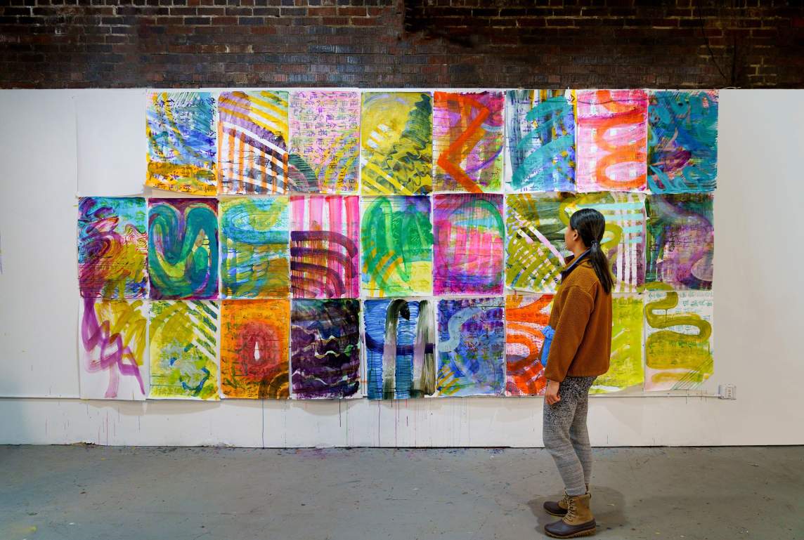 In an industrial room with white and brick walls and a concrete floor, a person with long black hair looks at a wall of brightly-colored, abstract artworks on paper. They are hung in a 9 x 3 grid on the white section of the wall, and include characters from a Chinese language in the background of the abstract compositions.