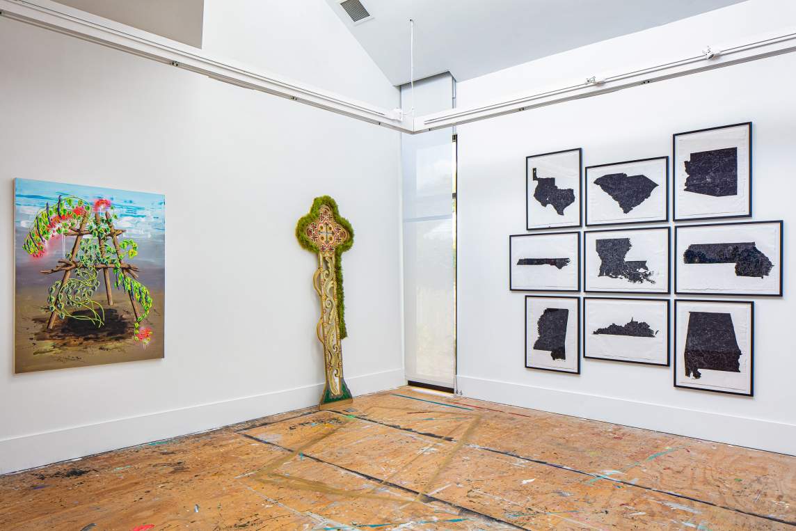 Three artworks hang in an art studio with white walls and distressed plywood floors. On the left is a painting with a wooden easel in an A shape, resting in the sand with the ocean behind it, covered over with vines and flowering plants. Next to it rests an ornate beaded and feathered sculpture in the form of a coptic cross.