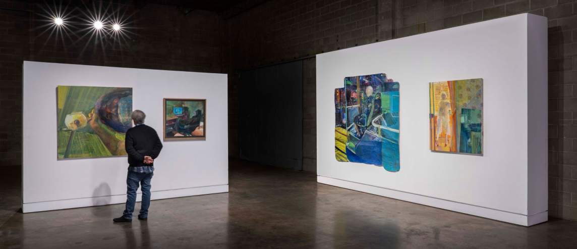 In an art gallery with floating white walls and a dark floor, a person with short hair stands with their back to the camera, looking at two paintings hanging on one of the walls. On the right side of the photo, is another section of wall with two brightly colored paintings hanging.