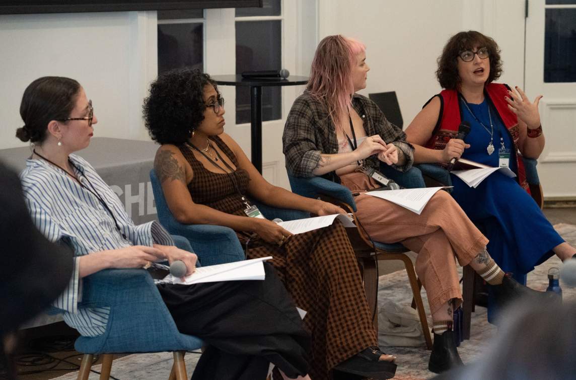 In blue chairs at the front of the room, 4 women sit in a panel discussion, all looking towards Sara Madandar on the far right who speaks into a microphone.