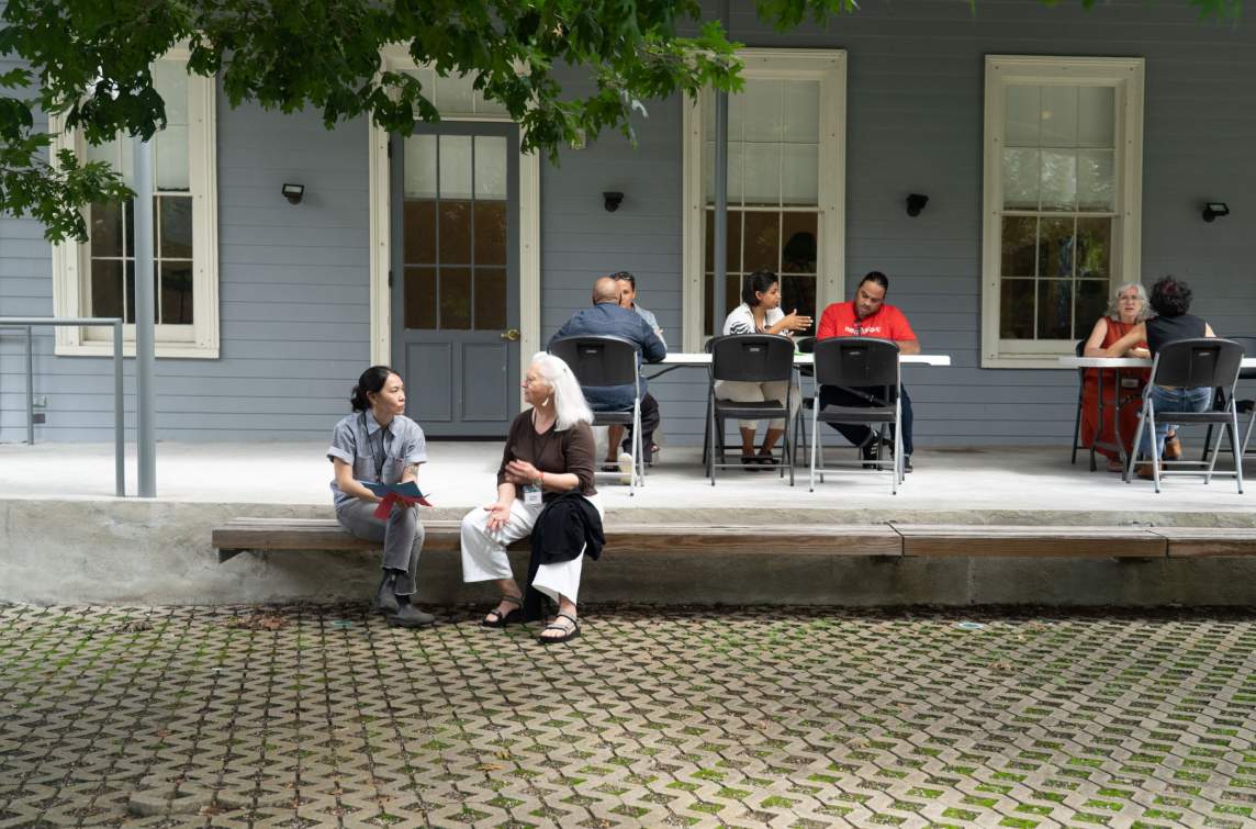 On a patio in front of a building with large windows and blue siding, various pairs of artists sit in discussion at tables and on a bench.