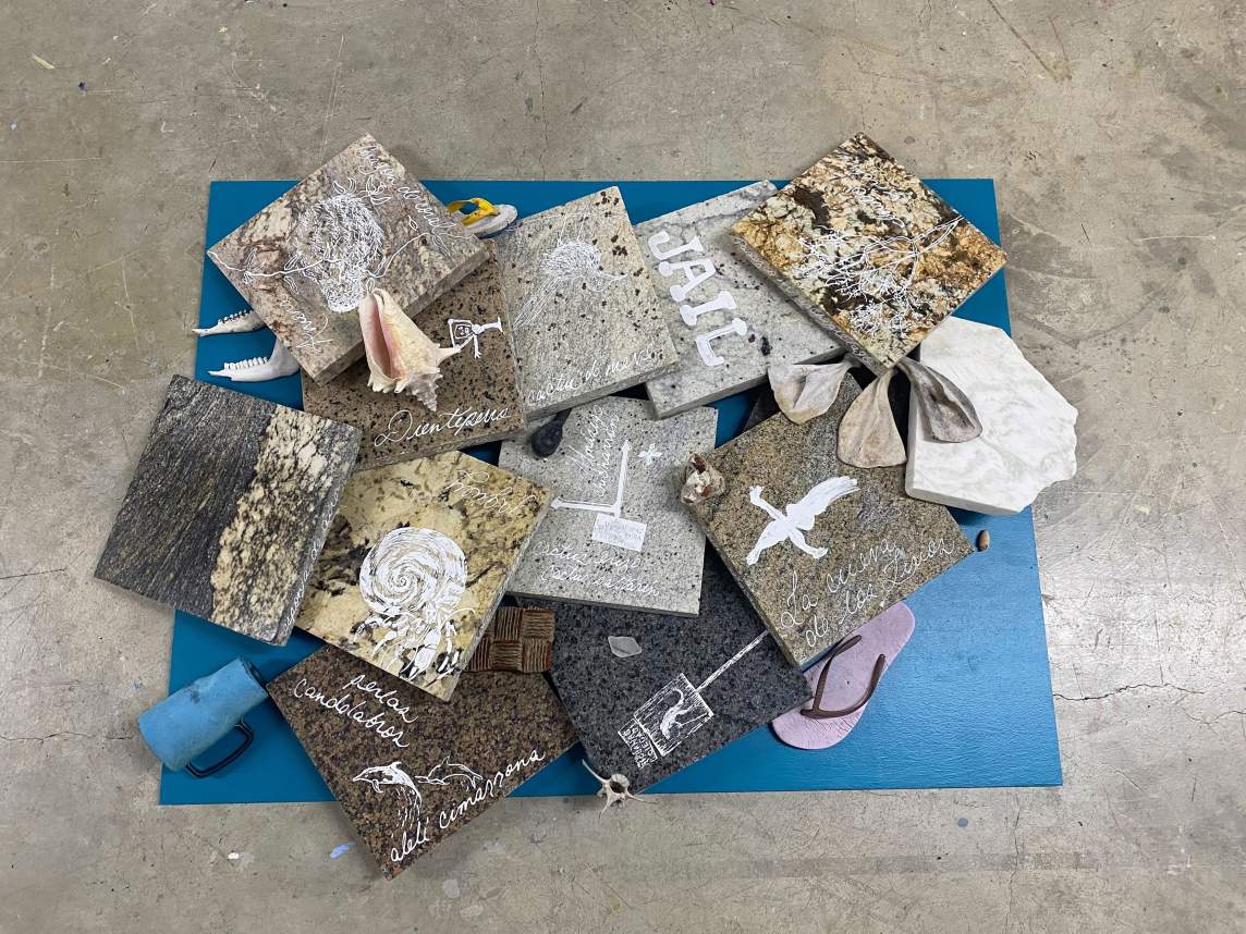 Photographed from above, a pile of nearly a dozen square slabs of polished granite lay on a blue yoga mat with flip flops, a travel cup, bones, seashells and other debris strewn amongst the slabs. The slabs have Spanish phrases and drawings on them, rendered in chalky white liquid paper, including drawings on shells, birds, and dolphins.