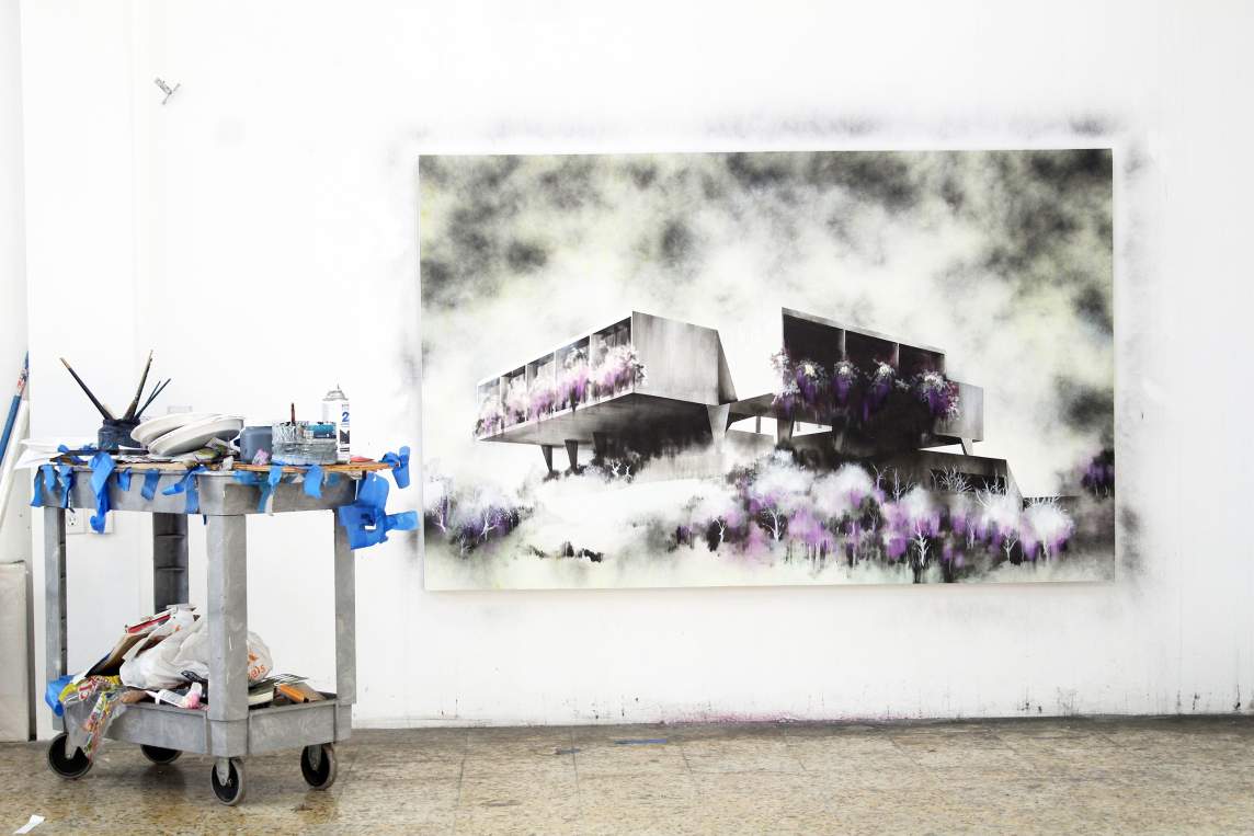 Painting of brutalist modern buildings with purple foliage on a white wall; art supplies on a cart to the left.