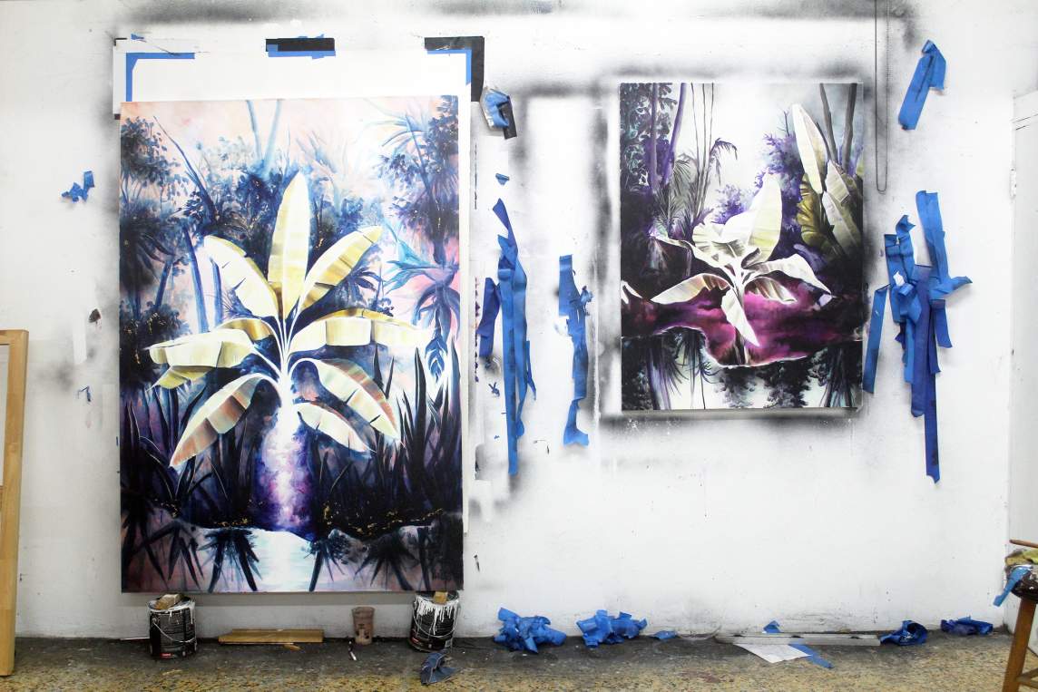 Painting of brutalist modern buildings with purple foliage on a white wall; art supplies on a cart to the left.