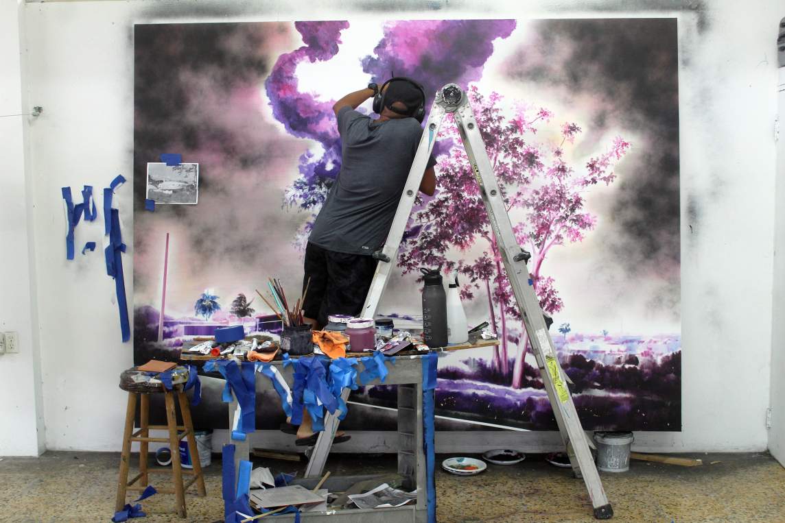 Artist on ladder paints a large landscape with purple smoke and trees on canvas. Cart below holds paint supplies, brushes, and tape.